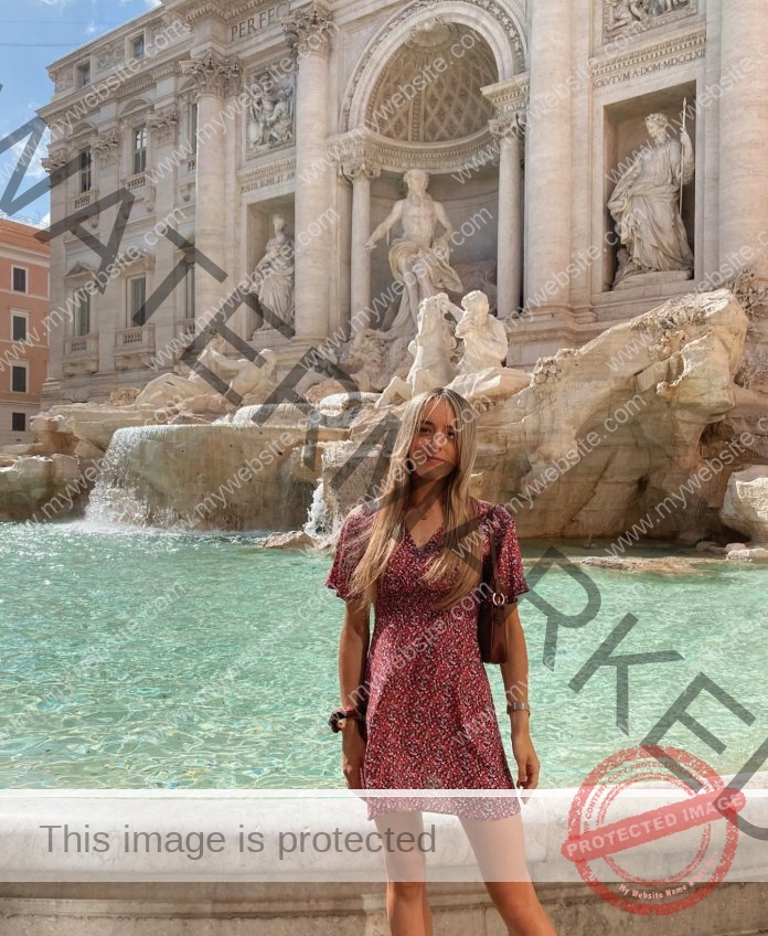 Judith Otazua, in a red patterned dress, stands before Rome's Trevi Fountain with turquoise water and sunlit marble statues behind her.