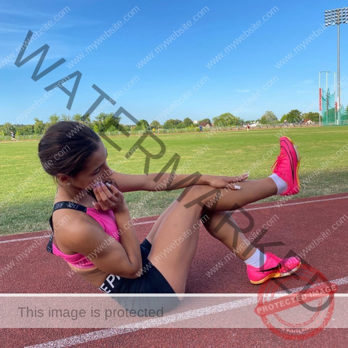 Iljana Beqiri in athletic wear does a bicycle crunch on a red track in Albania, grassy field and trees behind, bright pink shoes, pink bra.