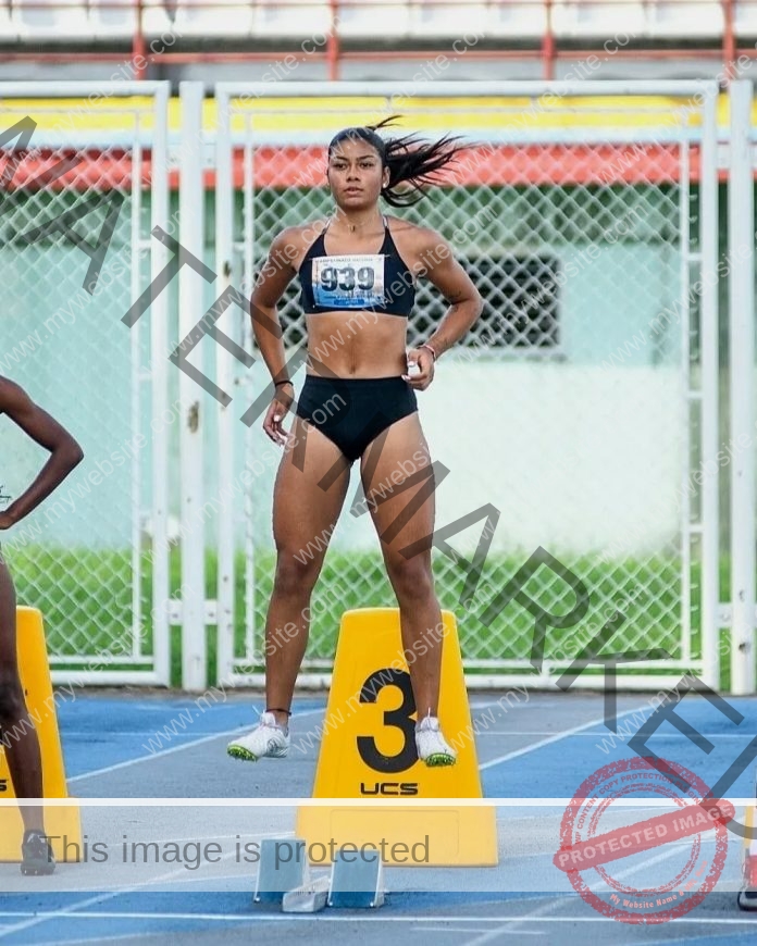 Orihana Guzman, in black sportswear, jumps behind lane marker 3 on a blue track, focused as she prepares to race with others nearby.