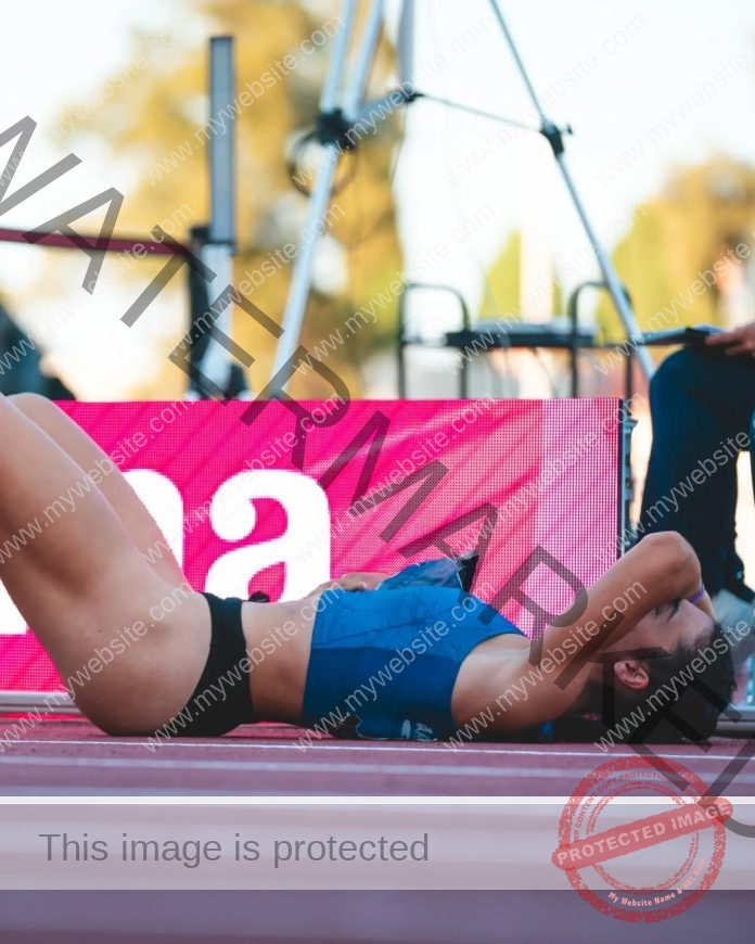 Jasynta Lampret lies on a running track in sportswear, covering her face with her hands after a race; pink sign in the background.