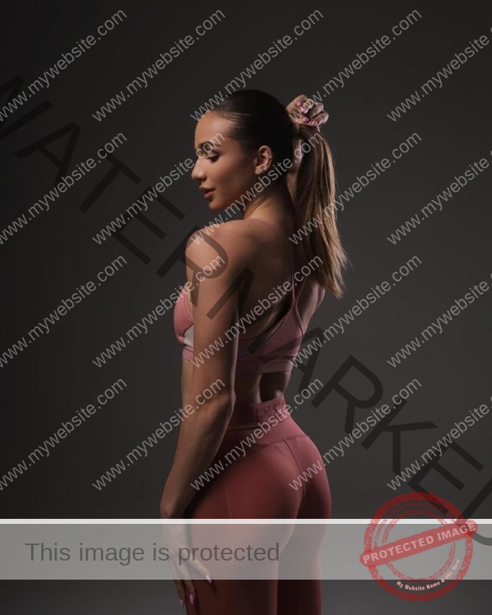 Siyana Brambarova posing in a darkened room wearing a light pink workout outfit and pulling up her ponytail.