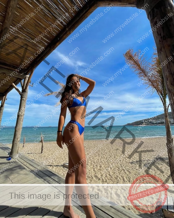Sofia Iosifidou, track and field star from Greece, stands on a beach walkway in a blue bikini, smiling with one hand to her head.