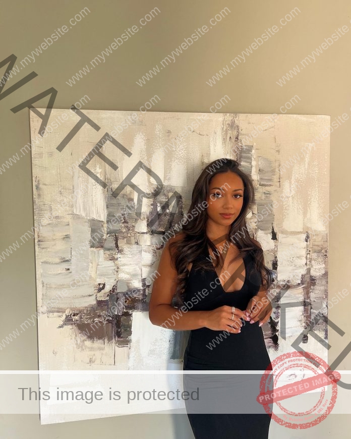 Janna Doumpas, with long wavy brown hair in a sleeveless black dress, stands by an abstract painting, looking at the camera neutrally.