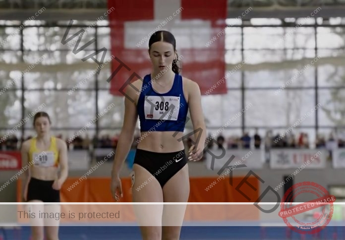 Xenia Buri, in a blue sports bra and black shorts with number 308, walks an indoor track; a Swiss flag hangs behind another athlete.