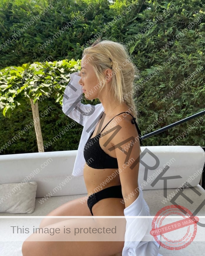 Xhuda Kruti with long blonde hair sits sideways on an outdoor sofa, in a black bikini and white shirt, relaxing after volleyball in Albania.