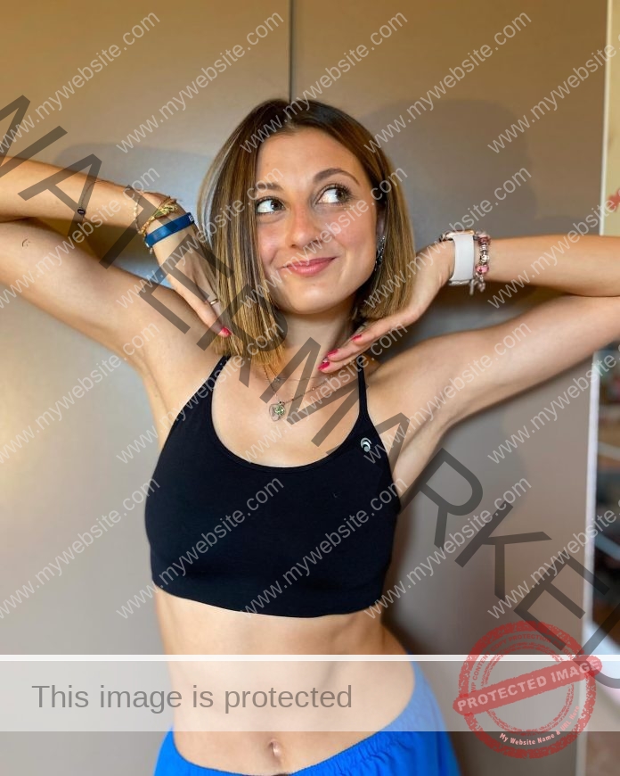 Idea Peroni in a flirty pose with hands under chin and smiling, wearing a black sports bra and flaunting her muscled midriff.