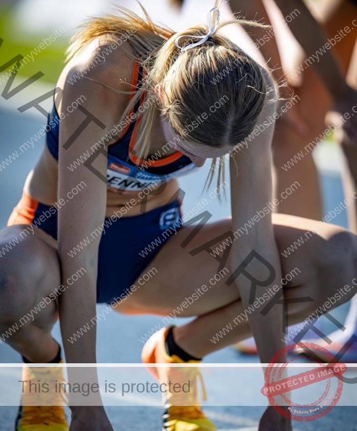 Alesha Bennetts crouches at the starting line in navy and orange, focused, sunlight on her face, with a white hair tie and yellow shoes.