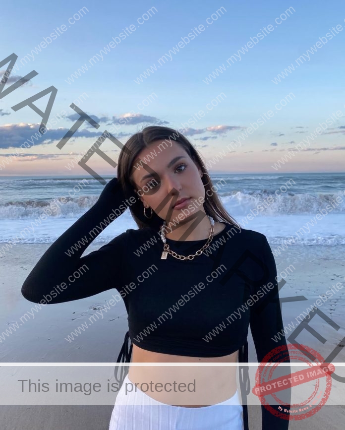 Agáta Cellerová standing on the beach, running her fingers through her hair, wearing a black tight long-sleeved shirt and white shorts.