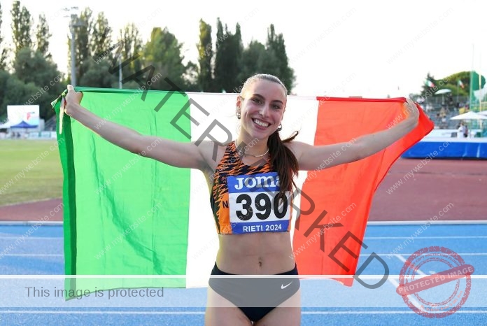 Agnese Musica spreads her arms wide holding the Italian flag behind her back with a big smile and an orange and black track uniform.