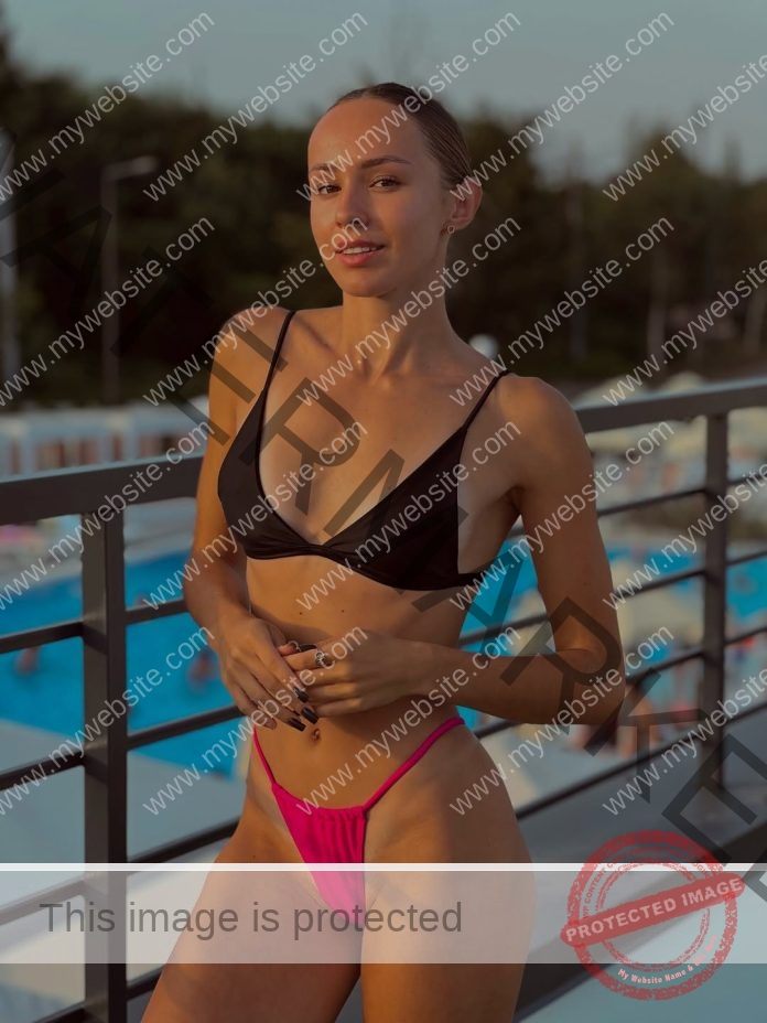 Alina Listunova standing near a pool, wearing a black bikini top and peach colored bottoms, smiling at the camera.