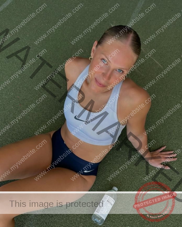 Annika Haldbo sits on green artificial turf in athletic wear, looking up at the camera with a water bottle nearby.