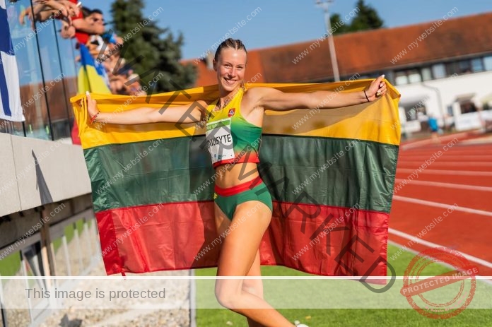 Aureja Beniusyte standing trackside, posing in her track uniform holding the Lithuanian flag behind her.