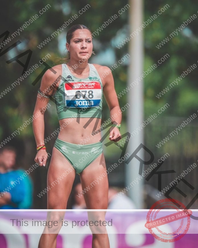 Carla Cruz Benlloch, a Spanish track athlete, stands outdoors in a green uniform with bib 978 and Joma shorts, crowd blurred behind.