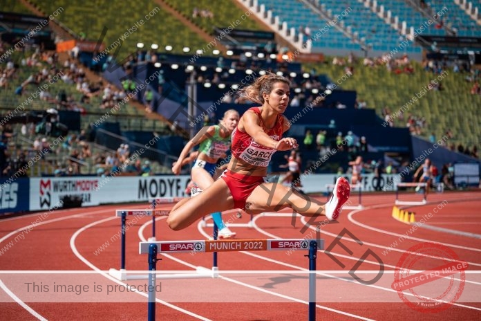 Carla García Sánchez clears a hurdle mid-race, showing speed and focus at a sunny stadium.
