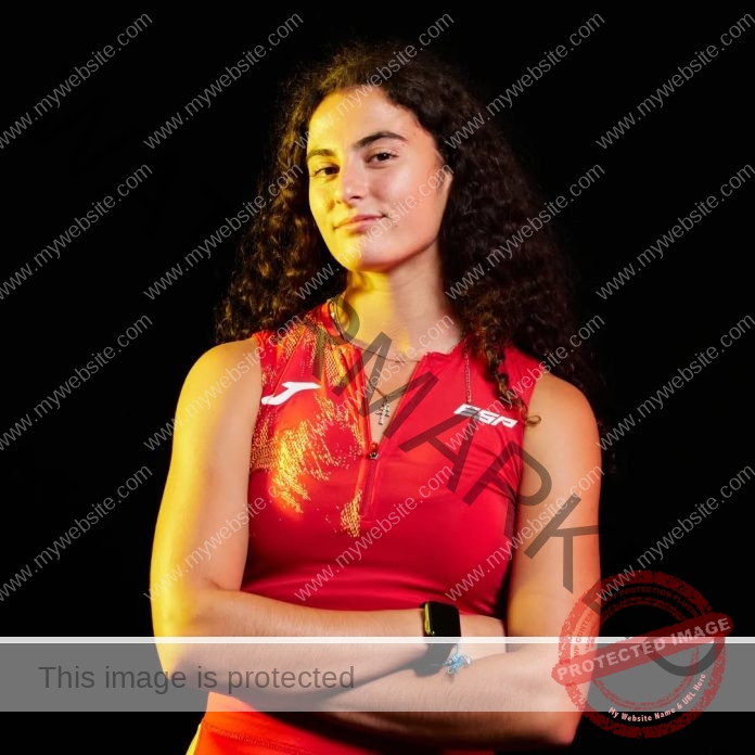 Carmen Marco Mora posing for a photo shoot, wearing a bright red athletic top, arms folded and smirking.