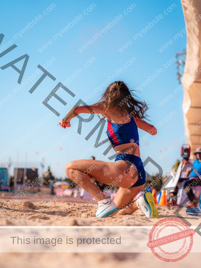 Daniela Rubio Pastor competing in a long jump event, mid-air over the sunny sand pit.