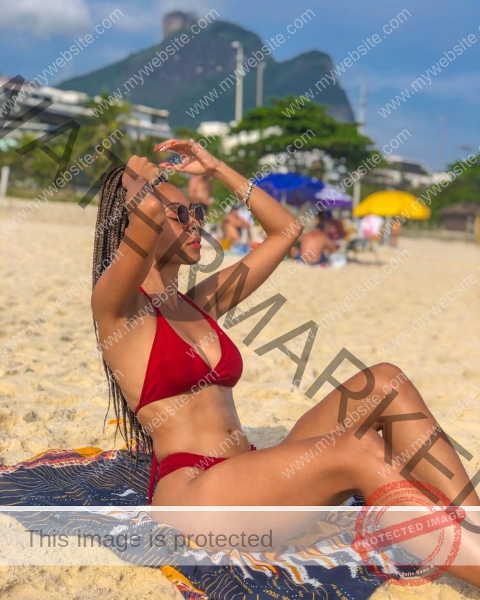 Ester dos Santos sitting on the beach in a red bikini, fixing her braided hair.
