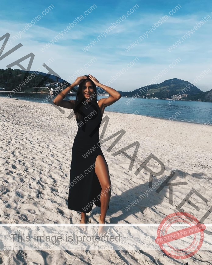 Julia Ribeiro posing on a sunny beach in a flowing black dress with scenic hills behind.