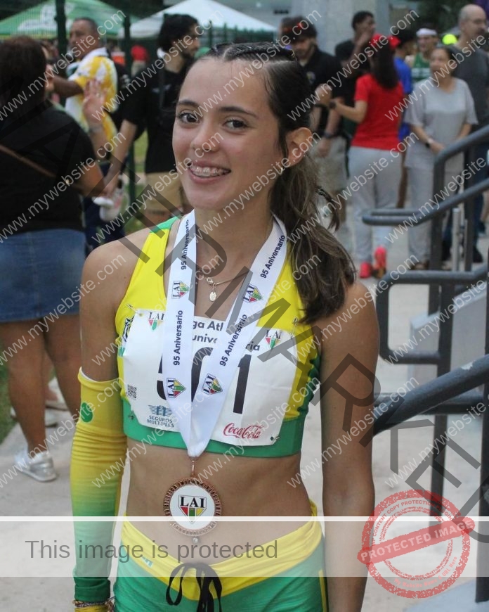 Legna Echevarria, a young female athlete in yellow and green, smiles with a medal outdoors near a fence in Puerto Rico.