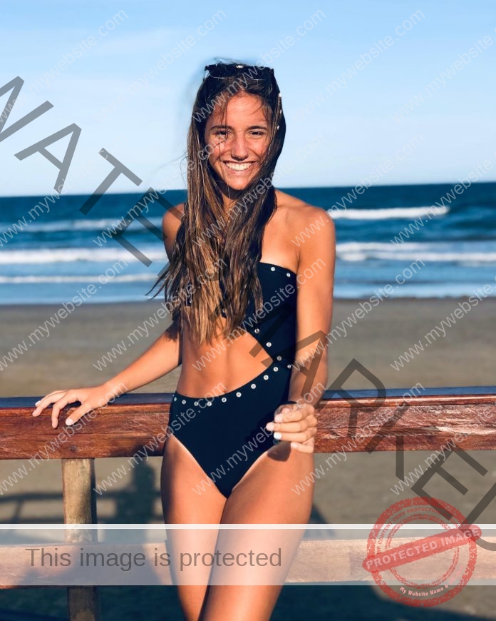 Lucia Gomez Irondo smiling by the ocean in a black swimsuit on a sunny beach day.