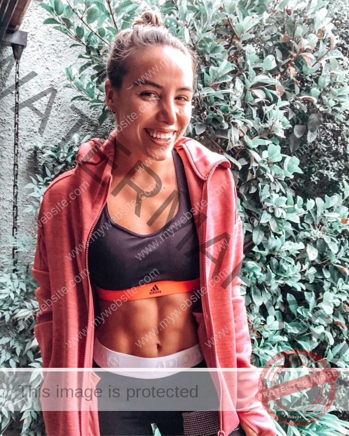 Macarena Borie smiles in black athletic wear and coral hoodie against green foliage outdoors.
