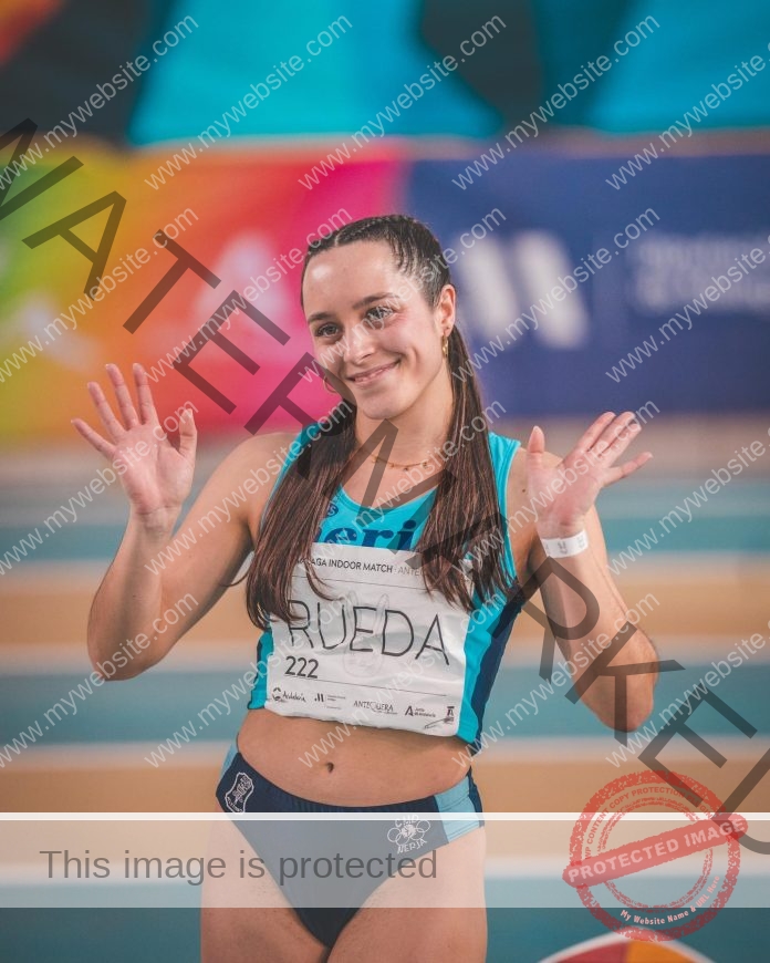 Maria Rueda waves to the crowd with both hands as she's introduced for a relay race wearing her baby blue track top and dark blue bottoms.