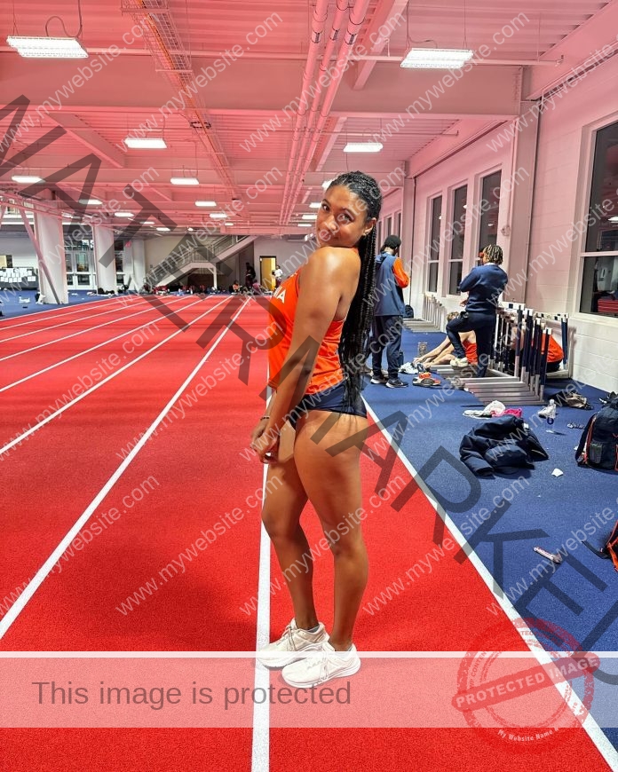 Maya Rollins standing on an indoor red track in orange top and black shorts.