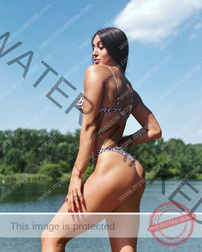 Mayya Gudanok poses by a Russian lake in a leopard-print bikini, looking over her shoulder with trees and blue sky behind her.