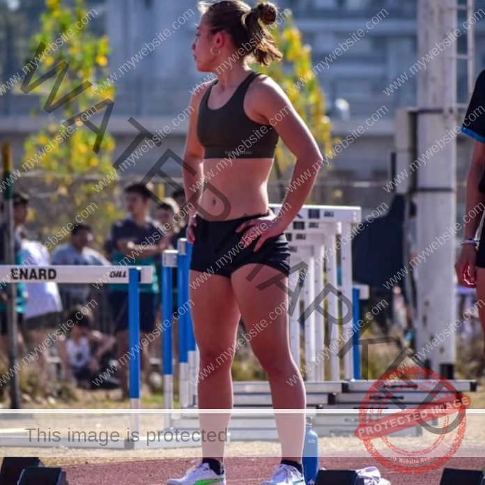 Paz Francucci, a female athlete in sportswear, stands on an outdoor track near hurdles, looking to the side with hands on her hips. Other people and equipment are visible in the background.