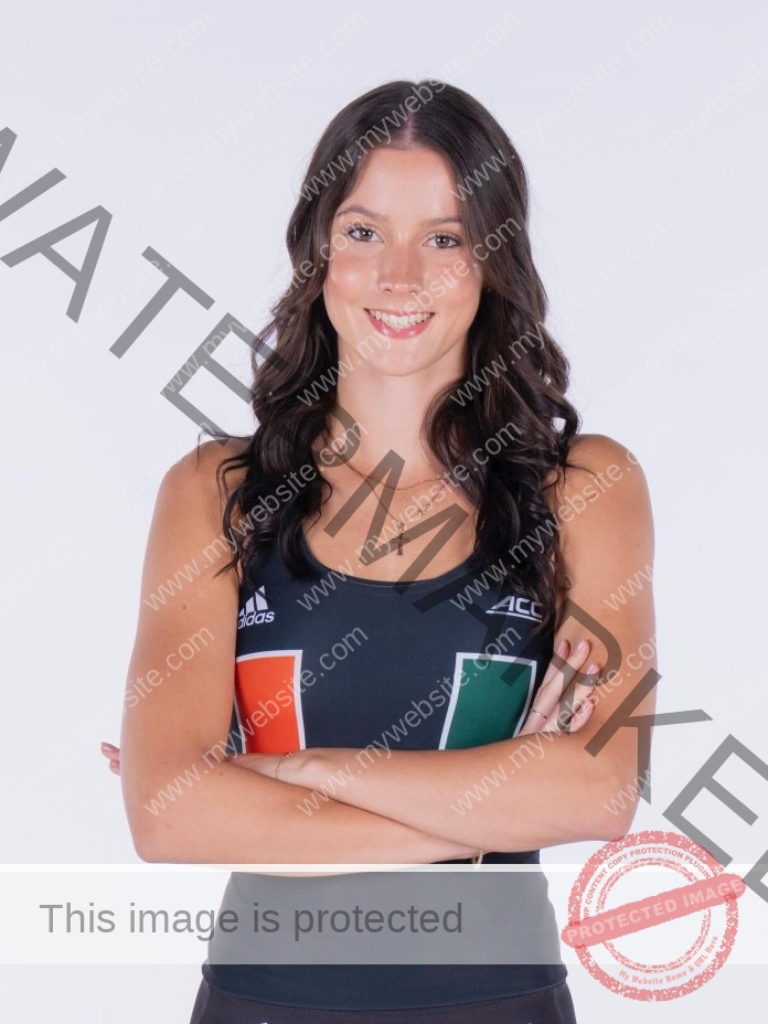 Adriana Kruzmane with long dark hair stands smiling, arms crossed, in a black sleeveless University of Miami top, white background.