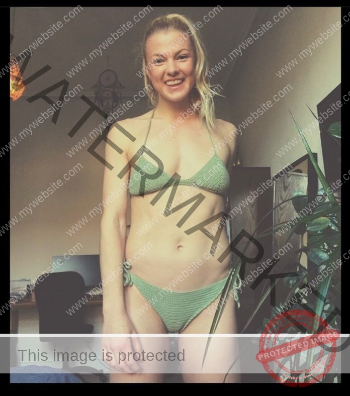 Anne Sofi Hyldtoft smiles in a green bikini indoors by a leafy plant, with a desk, chair, and dreamcatcher in the background.