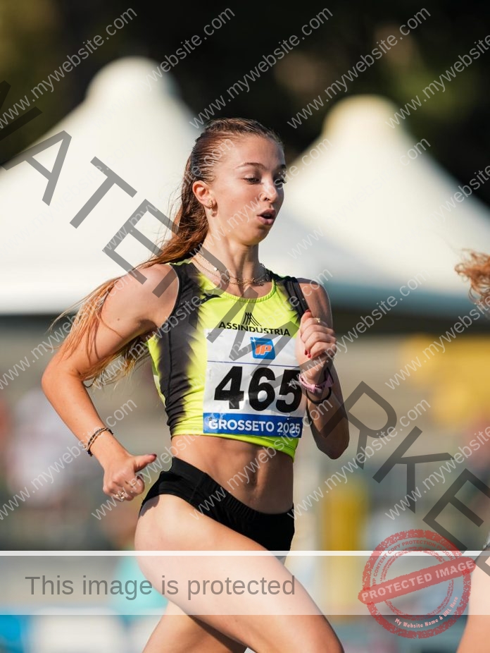 Giada Donati races in a yellow and black uniform with bib 465 marked "Grosseto 2025," focused as tents blur behind her.