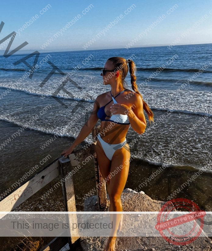 Karin Strametz in a blue and white bikini stands on a wooden platform by the ocean, looking to the side. The sun is shining, casting shadows, and gentle waves are visible in the background.