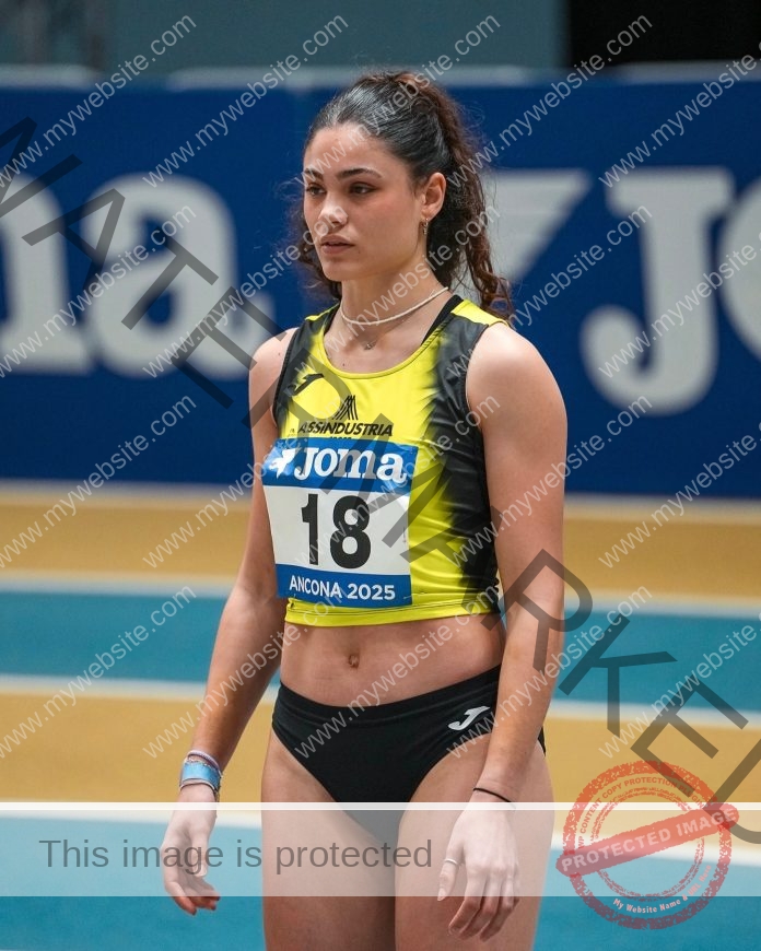 Laura Franceschi, in a yellow and black uniform numbered 18, stands focused on an indoor Italian track before her race; Joma banner behind.