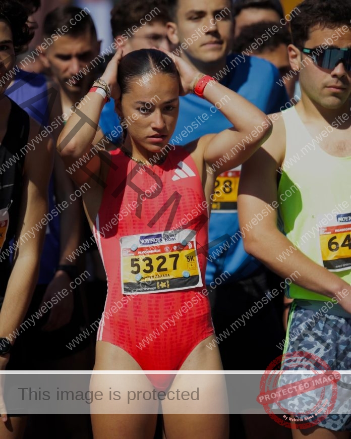 Myrto Valacha, a focused female track and field runner in red, adjusts her hair at the start line among other runners, bib 5327.