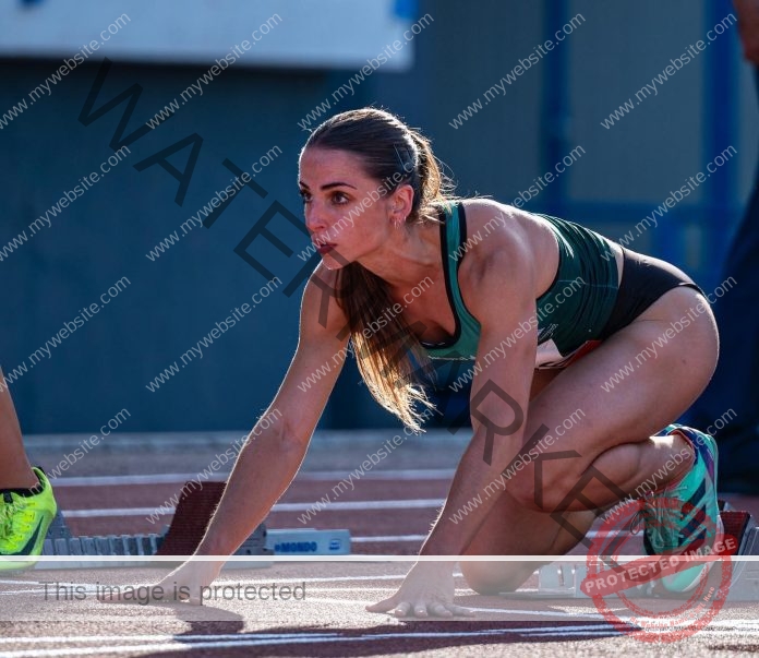 Leticia Arispe, a Bolivian track and field athlete in sportswear, crouches at the starting blocks on an outdoor track in sunlight.
