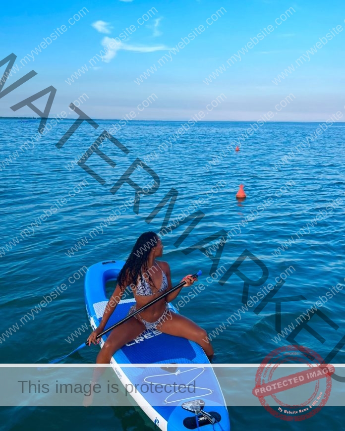 Rebecca Agbortabi sits on a blue paddleboard in calm water, holding a paddle. Two orange buoys float nearby under Italy's blue sky.