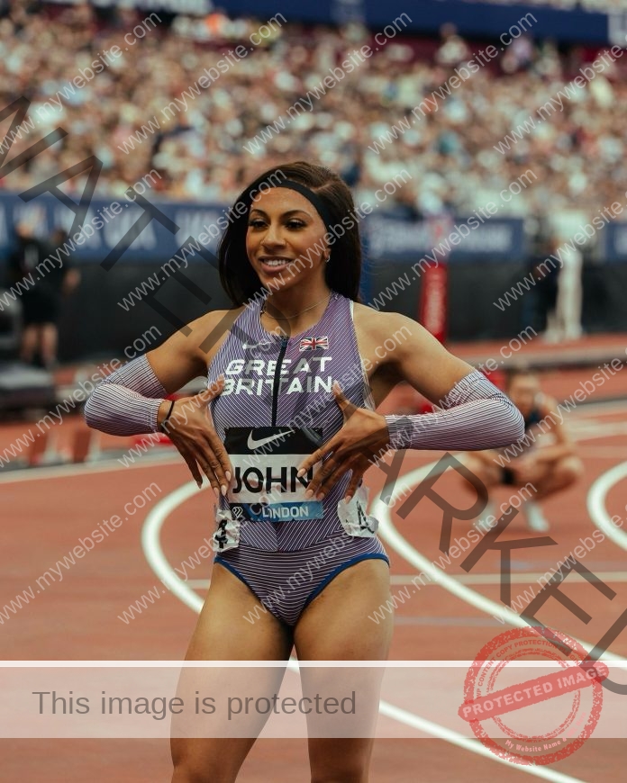 Yemi Mary John in a “Great Britain” uniform stands on a running track, smiling with hands on torso. UK stadium crowd behind her.