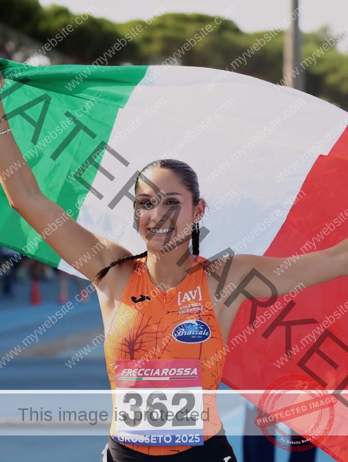 Zoe Tessarolo smiles, holding an Italian flag behind her, wearing orange and bib 362 on a track with trees in the background.