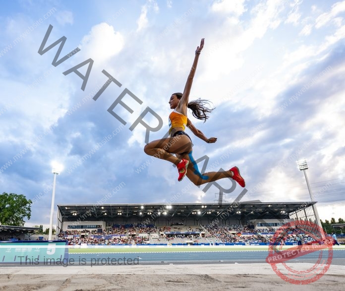 Carmen Rosales executing perfect long jump form in mid-air at track stadium competition.