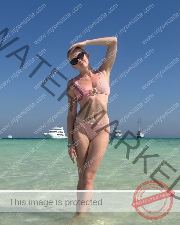 Iryna Baulina in a pink bikini stands in clear beach water, boats behind her, hand on head, sunglasses on, enjoying a training break.