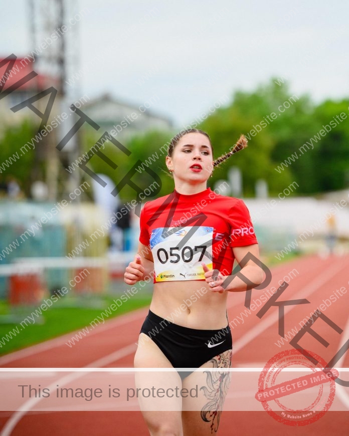 Anna Karundakova, in a red crop top and black shorts, races on an outdoor track with a braid and thigh tattoo. Blurred trees behind.