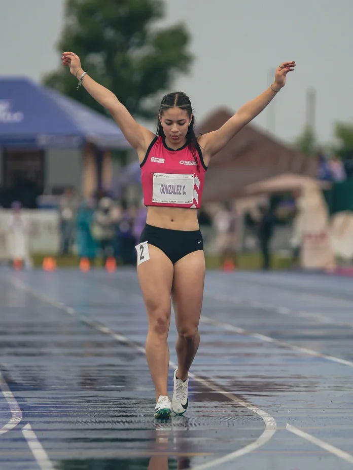 Camila Gonzalez, a female track athlete from Mexico, raises her arms crossing a wet finish line in pink and black with number 2.