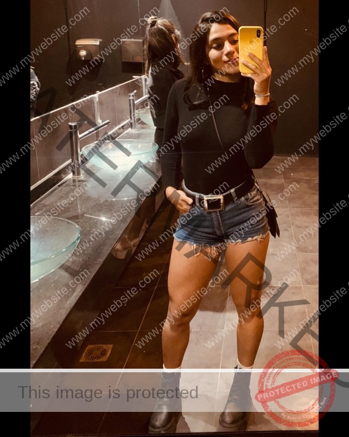 Camila Rodrigues takes a mirror selfie in a modern Mexico restroom, wearing a black top, denim shorts, boots, and white socks.