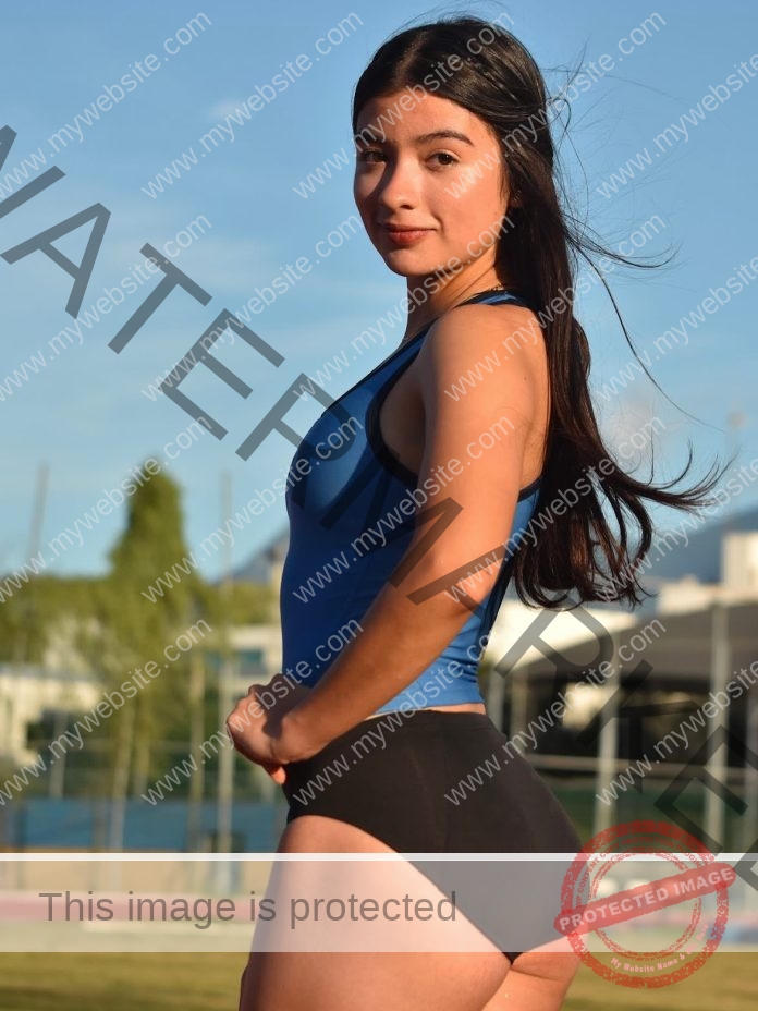 Dilean Flores poses outdoors in a blue tank top and black shorts, long hair flowing, green grass and buildings blurred behind her.