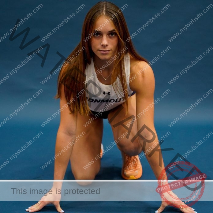 Emma Mckee in a Monmouth uniform crouches at the start on a blue track, looking determined, with long brown hair and orange shoes.