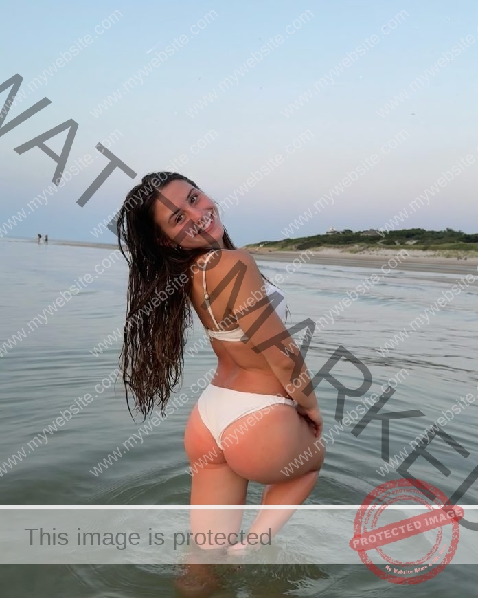 Hailey Denucci in a white swimwear standing in shallow beach water at sunset.