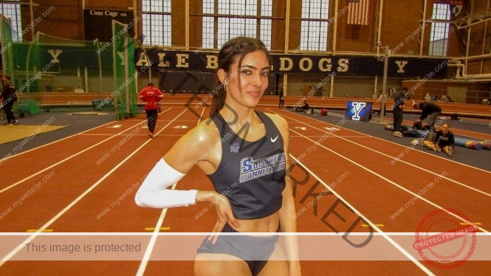 Hannah Caiola stands on an indoor track, smiling with one hand on her hip in a navy uniform and arm sleeve. YALE BULLDOGS behind.