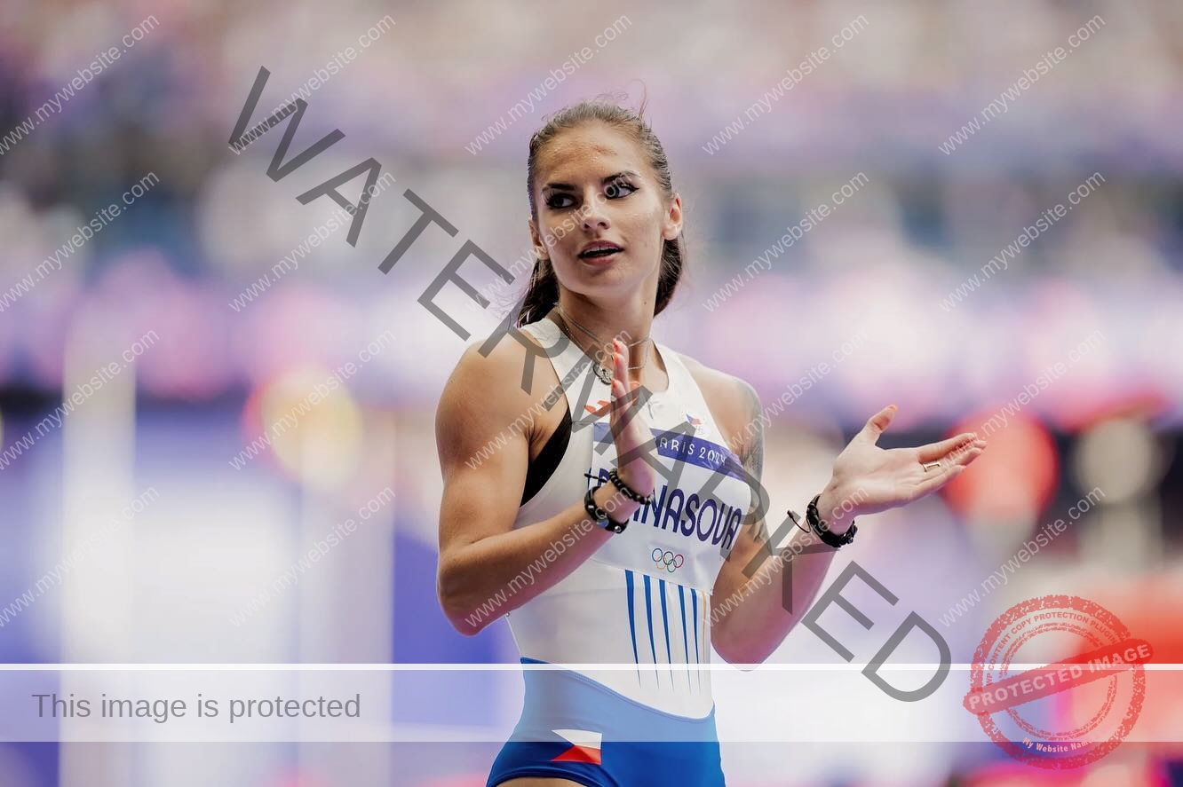 karolina-manasova-czechia-karolina_manasova-361 Karolína Maňasová, in a white and blue track uniform, stands clapping with hands raised, looking aside in a blurred indoor stadium.