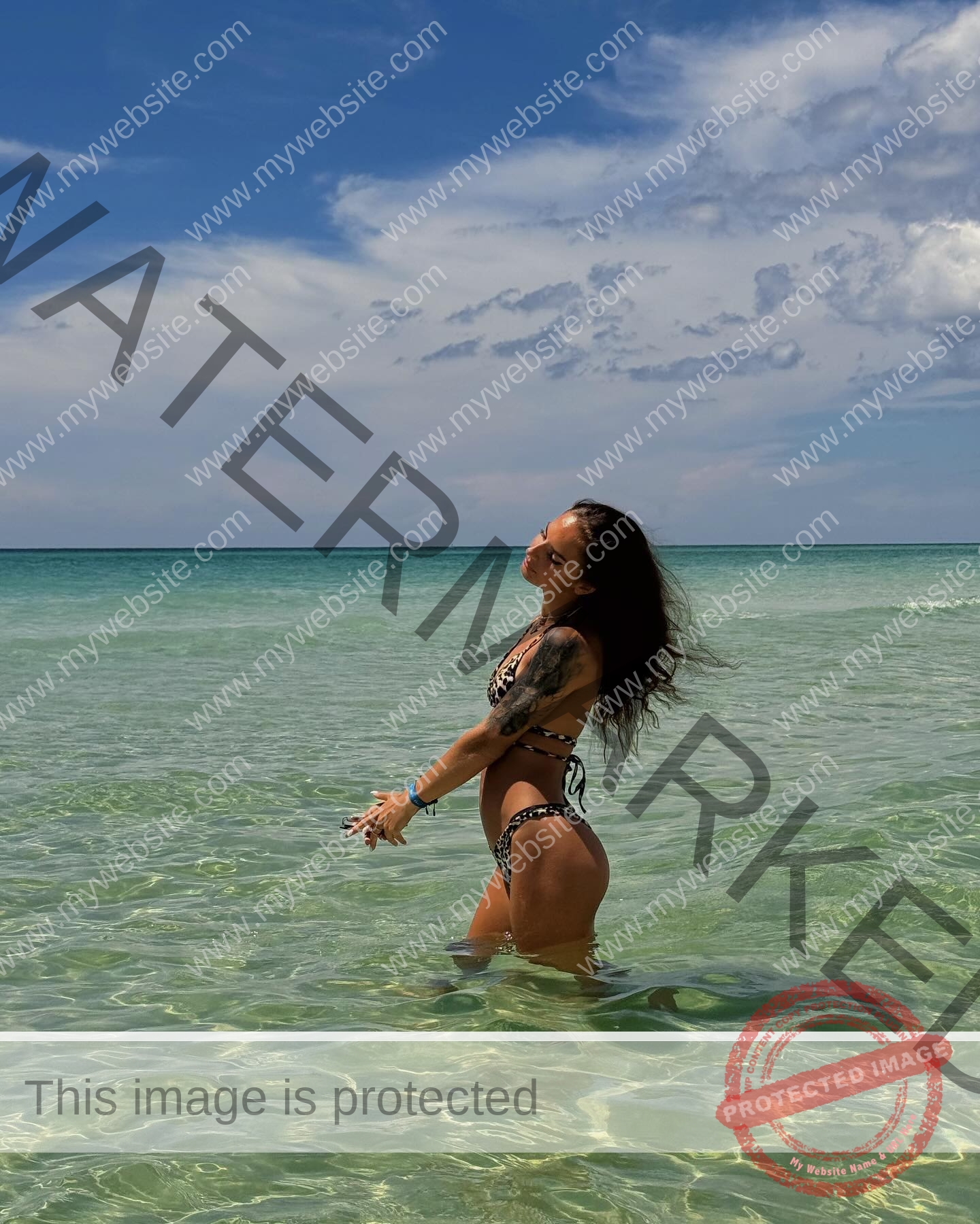 karolina-manasova-czechia-karolina_manasova-363 Karolína Maňasová in a patterned bikini stands in clear, shallow ocean water under a partly cloudy sky, hair blowing back.
